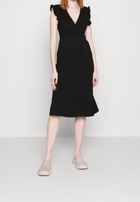 Robe portefeuille noire à manches volantées, longueur aux genoux. Texture lisse et coupe ajustée, associée à des chaussures beiges sans lacets.