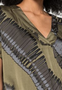 Grön silkesblus med v-ringning och en bred, flytande form. Svarta och grå abstrakta mönster pryder tyget. Slät textur.