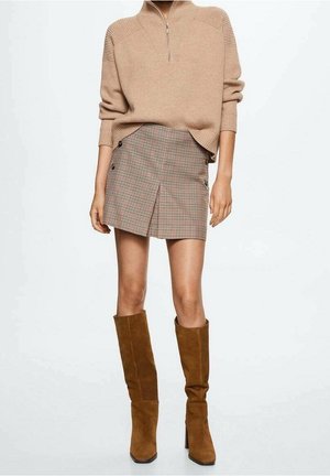 Person iført en beige lynlås-sweater, ternet mini nederdel med knapdetalje og brun ruskindsstøvler til knæet, stående.