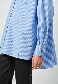 Light blue striped shirt with long sleeves, embellished with metallic floral accents and a single chest pocket; worn with black trousers.