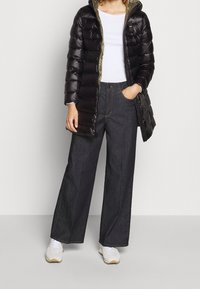 Kobieta ubrana w czarną kurtkę puchową, białą koszulkę, szerokie ciemne dżinsy i białe trampki, stojąca na tle jasnego, jednolitego tła.