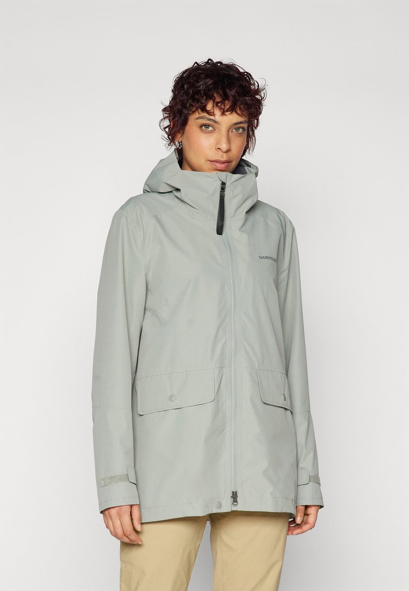 Veste imperméable gris clair avec capuche, fermeture éclair à l'avant et deux grandes poches appliquées. Présente une texture lisse et un design minimaliste.