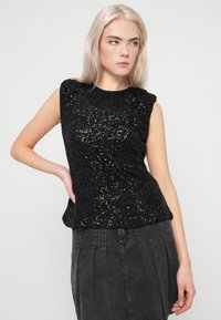 Mulher loira vestindo um top preto de lantejoulas sem mangas e uma saia de ganga escura, posando com uma mão na anca, olhando para o lado em fundo liso.