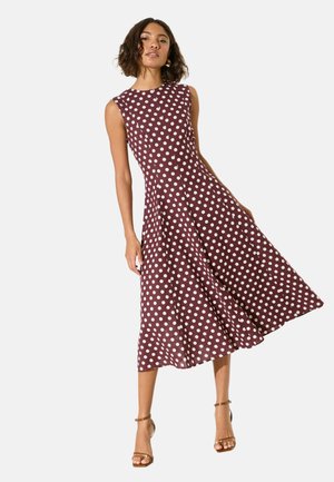 Moteris dėvi rankovių neturintį bordo midi ilgio suknelę su baltais taškais ir rudos spalvos kulnus su kulkšnies dirželiais, stovinti priešais baltą foną.