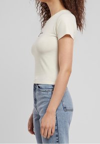 Krämfärgad t-shirt med kort ärm och en figurnära design, slät textur och liten svart tryck på framsidan, matchad med högmidjade blå denimjeans.