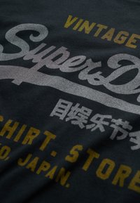 Zwart katoenen T-shirt met de tekst "Super Dry" in een zilver- en goudverloop. Bevat Japanse karakters en een label met "VINTAGE". Gladde textuur.