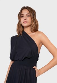 Femme aux cheveux châtain clair ondulés portant une robe noire à une épaule, la main sur la hanche, sur un fond gris clair uni.