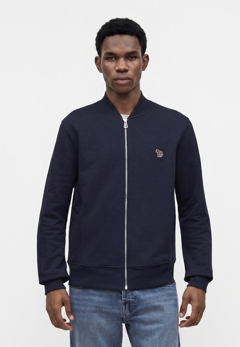 Homme portant une veste zippée bleu marine avec un petit emblème zèbre sur la poitrine et un jean bleu, debout devant un fond blanc uni.