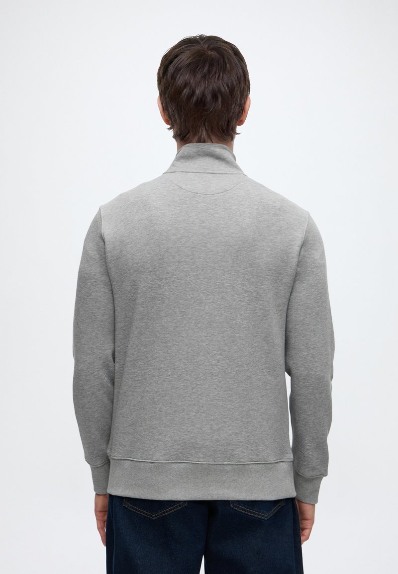 Mann mit dem Rücken zur Kamera, trägt einen hellgrauen Pullover und dunkelblaue Jeans vor einem schlichten weißen Hintergrund.