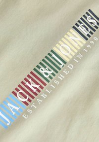 Έγχρωμο ριγέ κείμενο που γράφει «Jack & Jones» με το «Ιδρύθηκε το 1990» από κάτω, σε ανοιχτό μπεζ υφασμάτινο φόντο.