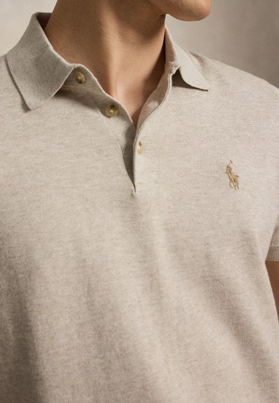 Κοντινό πλάνο ενός ατόμου που φοράει ένα μπεζ πλεκτό πόλο με δύο ξεκούμπωτα κουμπιά και ένα μικρό κεντητό λογότυπο αλόγου στο στήθος.