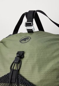 Mochila verde oliva con un patrón geométrico, con correas negras y un logotipo. Incluye clips ajustables y un detalle de bolsillo de malla.