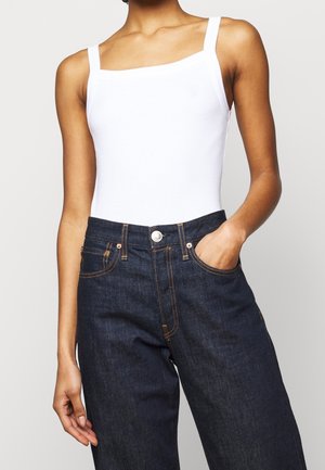 Białe body z kwadratowym dekoltem w zestawieniu z ciemnoniebieskimi jeansami z wysokim stanem, posiadającymi przednią kieszeń i kontrastujące pomarańczowe przeszycia.