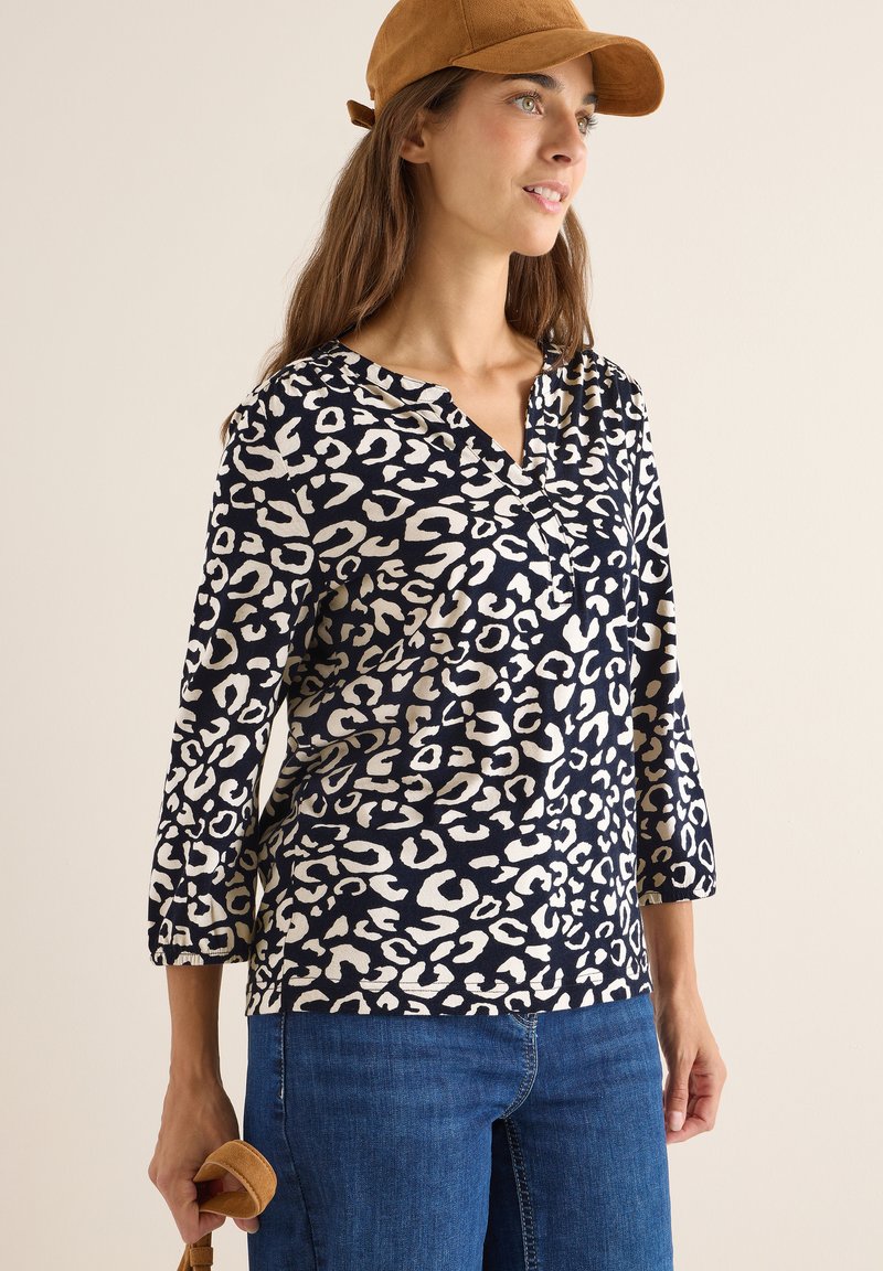 Top leopard a stampa navale con scollatura a V e maniche tre quarti, realizzato in tessuto morbido. Modella che tiene un accessorio beige, indossando un cappello beige.