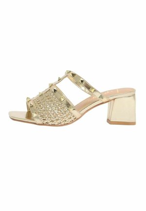 Goldfarbene Mule-Sandale mit offenem Zehenbereich, Blockabsatz, geflochtenem Vorderriemen und goldenen Pyramidennieten entlang mehrerer Riemen.