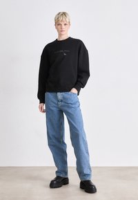 Svart hettegenser med "Calvin Klein Jeans" logo på brystet, kombinert med lyseblå denimjeans med løs passform og fyldige svarte plattformsko.