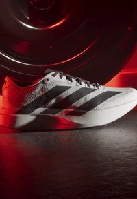 Weißer Laufschuh mit schwarzen Streifen und "Lightstrike Pro"-Sohle, auf einem Boden mit einer großen schwarzen Gewichtsscheibe in roter Beleuchtung platziert.