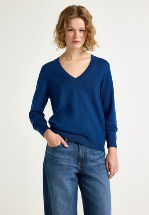 Donna con capelli ricci che indossa un maglione blu con scollo a V e jeans blu, in piedi con una mano in tasca su uno sfondo semplice.