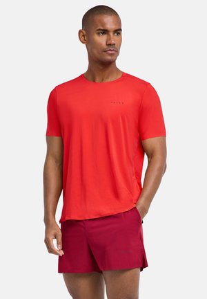 Man draagt een felrood T-shirt met korte mouwen en donkerrode shorts, staat met één hand in de zak en kijkt opzij tegen een lichte achtergrond.