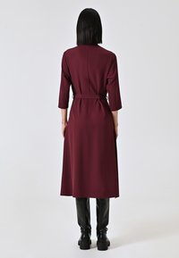 Robe bordeaux à manches longues avec une taille cintrée, détail de ceinture nouée et texture unie, associée à un pantalon noir ajusté et des bottes.