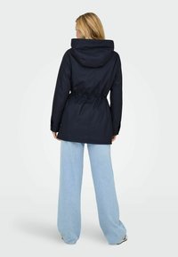 Veste imperméable bleu marine avec taille à cordon de serrage ajustable, capuche oversize et manches longues, associée à un pantalon large bleu clair.