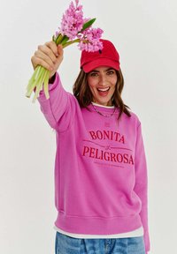 Ružový mikina s červeným textom "BONITA y PELIGROSA" nosená cez bielu košeľu. Model drží ružové kvety a má na hlave červenú čiapku.