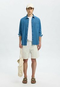 Chemise en jean bleue, t-shirt blanc, short beige à rayures, sandales vertes et casquette légère. Tient un sac fourre-tout beige. Tenue simple et décontractée.