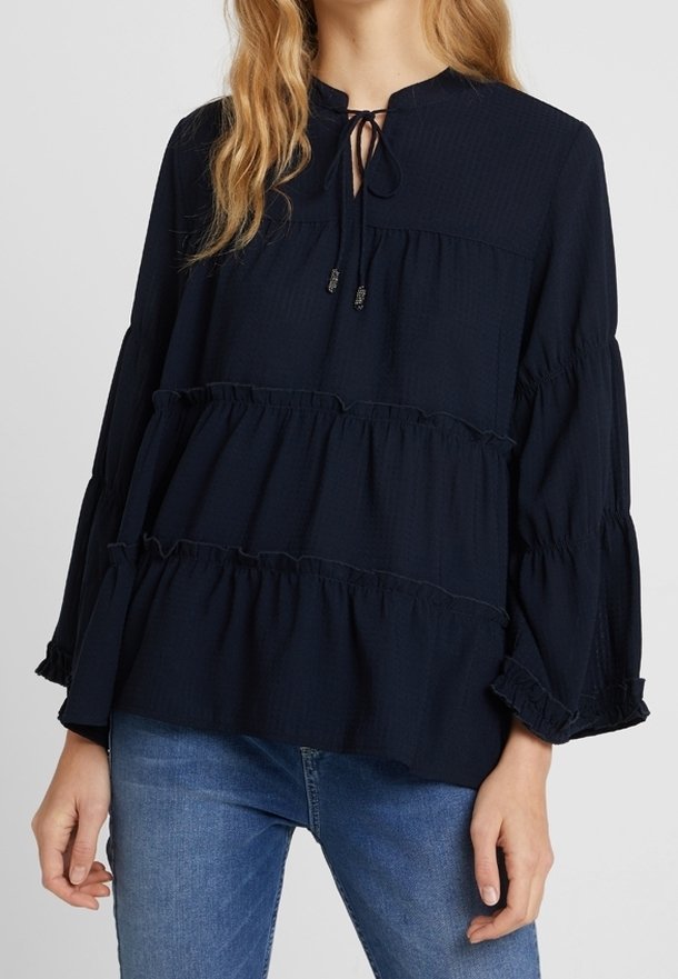 Femme portant un chemisier bleu marine ample à manches longues avec des volants superposés et une fermeture à nouer au niveau du col, assorti à un jean bleu.