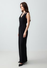 Zwarte jumpsuit met een V-halslijn, aansluitende bovenkant en wijde pijpen. Voorzien van een ceintuur in de taille en subtiele plooien, gecombineerd met zwarte puntige schoenen.