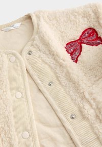 Giacca in peluche color crema con texture morbida, caratterizzata da un dettaglio a fiocco rosso in paillettes. Interno in corduroy beige chiaro e chiusura con bottoni a pressione.