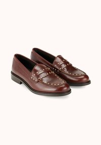 Mocassins en cuir marron avec des accents cloutés sur le dessus, un bord festonné décoratif et une semelle plate en caoutchouc. Design classique avec une finition polie.
