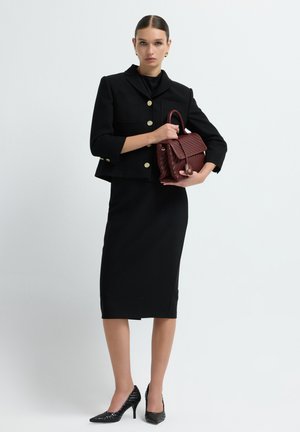Donna in tailleur nero con bottoni dorati, che tiene una borsa a tracolla di un rosso scuro testurizzato, indossando tacchi neri testurizzati, in piedi su uno sfondo bianco.