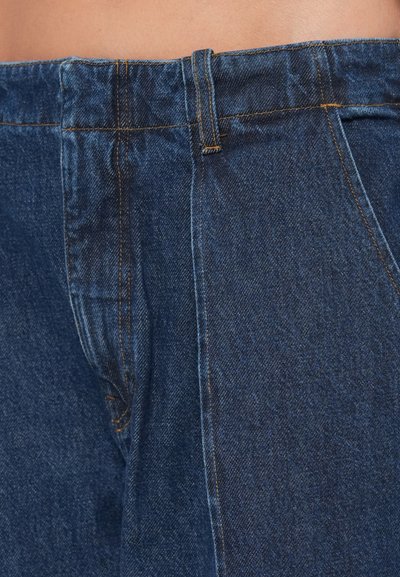 Közeli kép egy személyről, aki sötétkék farmernadrágot visel, látható varrással és övbújtatókkal deréktájon.