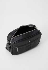 Svart skinn crossover-veske med sølvfarget detaljer, en glidelås hovedrom, og et teksturert frontpanel med en logo.