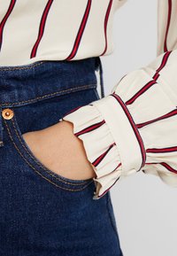 Blusa crema con rayas verticales rojas y marinas, con un puño con volantes. Combinada con jeans oscuros de tiro alto.