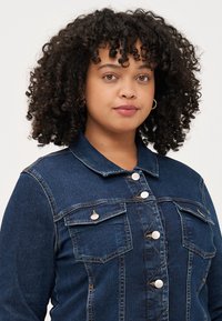 Mlada ženska z curled lasmi nosi jeans jakno z detajli gumbov, pred nevtralno ozadje. Z zaupanjem gleda proti opazovalcu.
