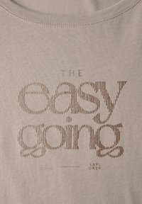 Camiseta de color beige claro con texto en relieve en bronce oscuro que dice "THE easy going" con un acabado texturizado y cuello de crew.