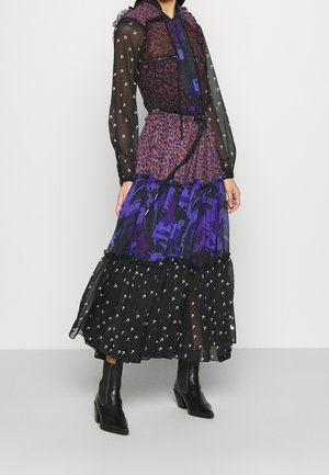 Žena v dlouhých vrstvených šatech s fialovým leopardím vzorem, modrými abstraktními motivy a černou průsvitnou látkou s bílými šipkami, doplněných černými botami.