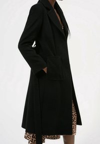 Personne portant un long manteau noir sur une robe à imprimé léopard, la main glissée dans la grande poche avant du manteau.