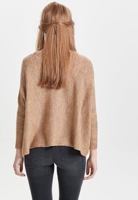 Béžový svetr z vlněné směsi s volným střihem, širokými rukávy a žebrovanými detaily. Zadní část je lehce delší než přední. Nosený s tmavými džíny.