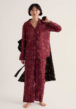 Femme debout pieds nus portant une chemise à manches longues et un pantalon assortis à motif floral rouge, tenant un manteau floral foncé sur son épaule.