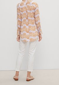 Blusa a maniche lunghe con un motivo di foglie multicolori in tonalità di arancione su uno sfondo chiaro, abbinata a pantaloni bianchi slim e sandali.