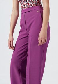 Lila skräddarsydda byxor i mjukt tyg; har hög midja, veck och en liten metallaccent nära fickan. Slät textur.