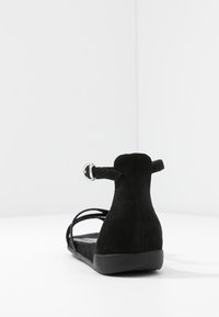 Schwarze Wildleder-Sandale mit flacher Sohle, offenem Rücken, mehreren dünnen Riemen und verstellbarem Knöchelriemen. Glatte Textur und minimalistisches Design.