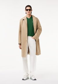 Cappotto lungo beige sopra una t-shirt verde, pantaloni bianchi e sneakers multicolori. Modello con occhiali da sole neri. Tessuto liscio e vestibilità comoda.