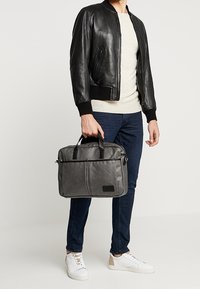 Sort læder bomberjakke, lys grå skjorte, blå jeans, grå laptop taske med sorte accenter og hvide sneakers. Simpel og funktionel design.