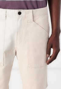 Bézs színű, pamut anyagú cargo short, gombos záródással, oldalsó zsebekkel és finom textúrával, részben látható kéz mellett bemutatva.