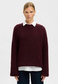 Burgunderfarbener Strickpullover mit lockerer Passform, überschnittenen Schultern und gerippten Bündchen. Über einem weißen Hemd mit Kragen getragen; weiche Textur.