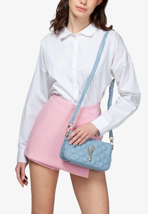 Donna con camicia bianca abbottonata e minigonna rosa, che tiene una borsa a tracolla trapuntata azzurro chiaro con logo e dettagli dorati.