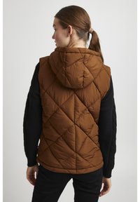 Gilet matelassé marron avec capuche, doté d'un revêtement en losanges angulaires. Associé à un haut noir texturé à manches longues et à un pantalon noir.
